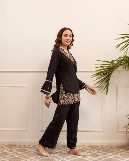 Woman wearing a black traditional outfit with floral embroidery in a room with a white wall and patterned floor.