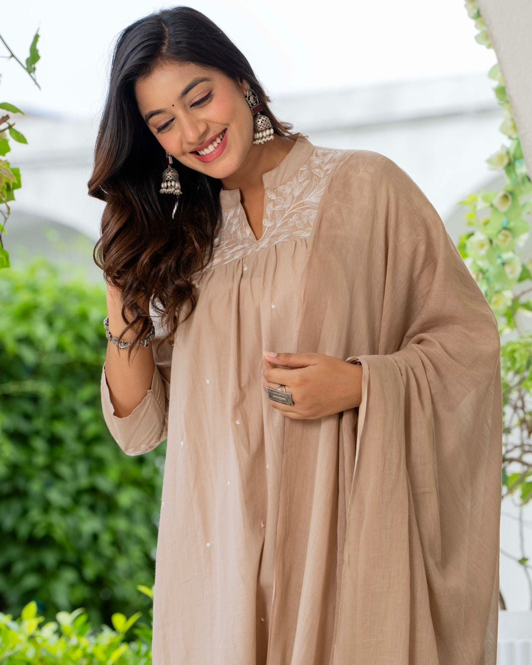 Woman wearing a beige dress with a floral pattern, standing outdoors with greenery in the background.