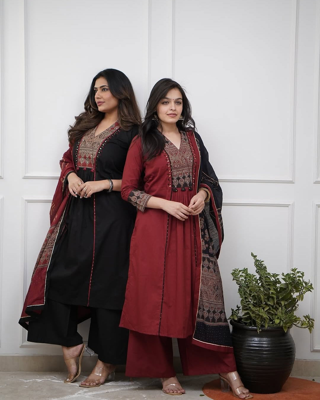 Two women in traditional outfits standing against a white paneled wall with a plant in the foreground.