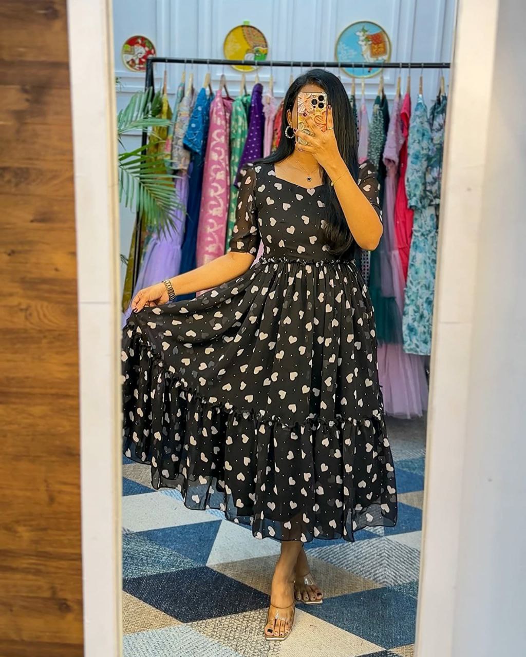 Woman in a black dress with white patterns taking a mirror selfie in a clothing store.