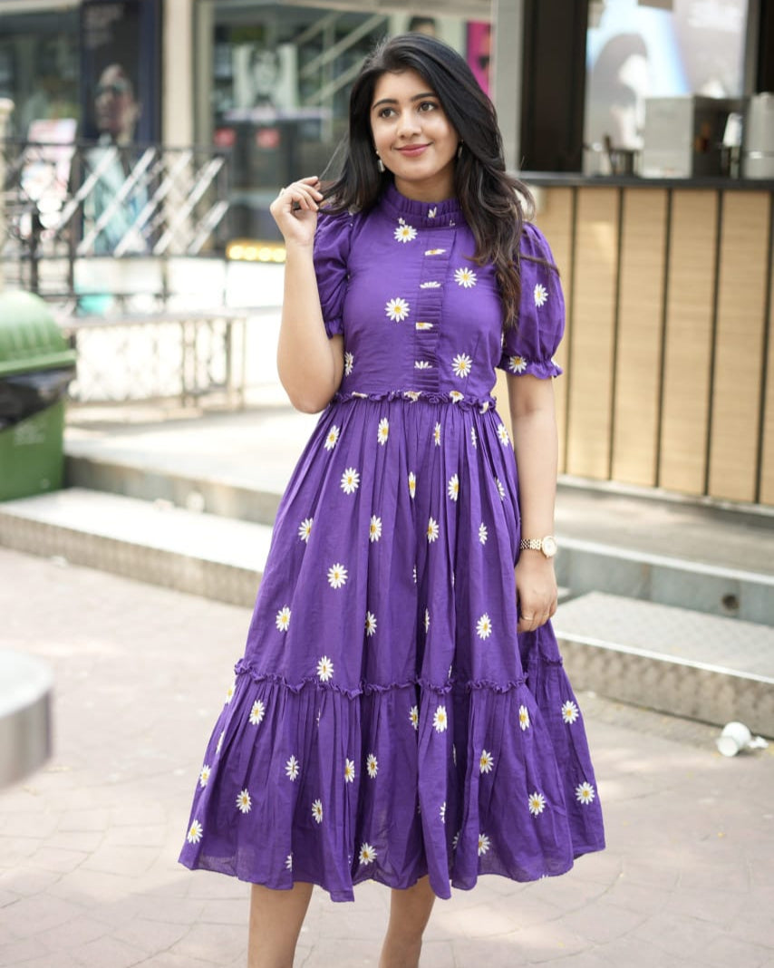 Woman wearing a purple dress with floral patterns standing outdoors.