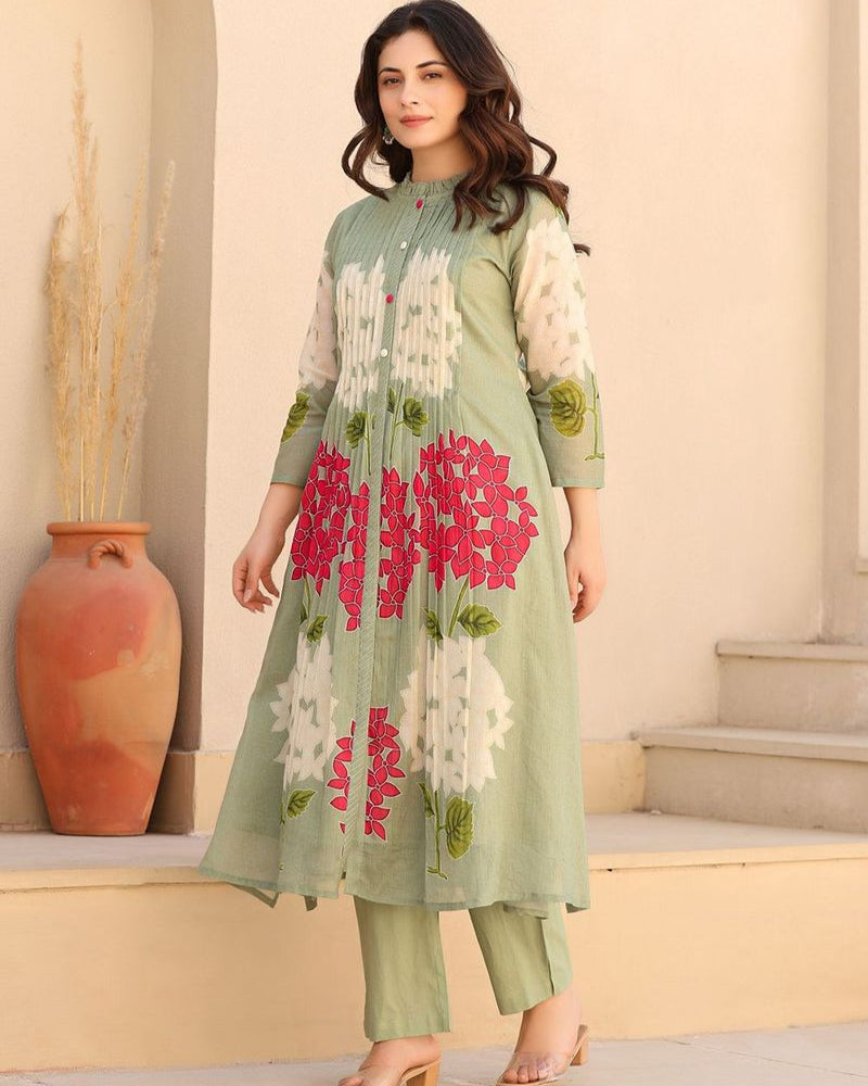 Woman wearing a light green floral dress standing outdoors with a beige wall and vase in the background.