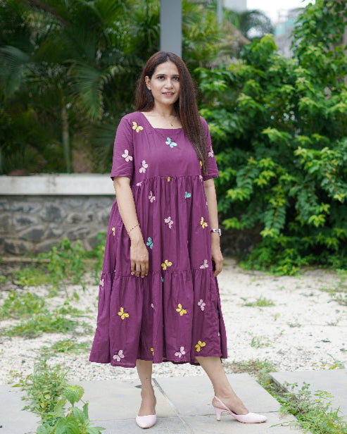 Woman in a purple dress with floral patterns standing outdoors on a path with greenery.