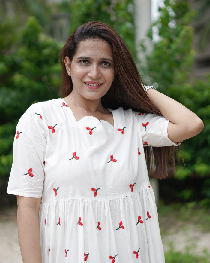 Woman in a white dress with red patterns standing outdoors with greenery in the background