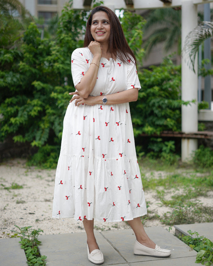 Woman in a white dress with red patterns standing outdoors on a sidewalk.