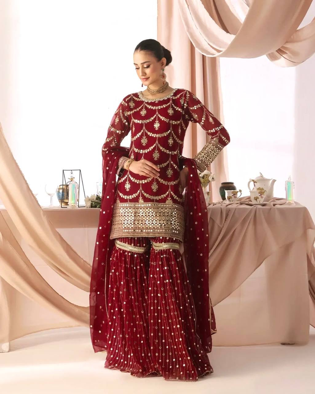 Woman in a red and gold traditional outfit standing in a decorated room with curtains and a table.