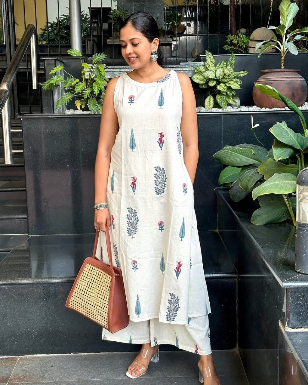 Woman in a white dress with floral patterns standing outdoors near plants.