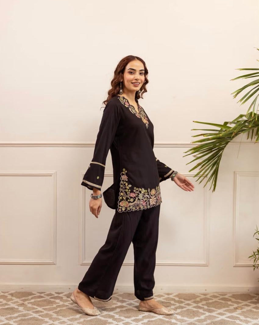 Woman wearing a black traditional outfit with floral embroidery in a room with a white wall and patterned floor.