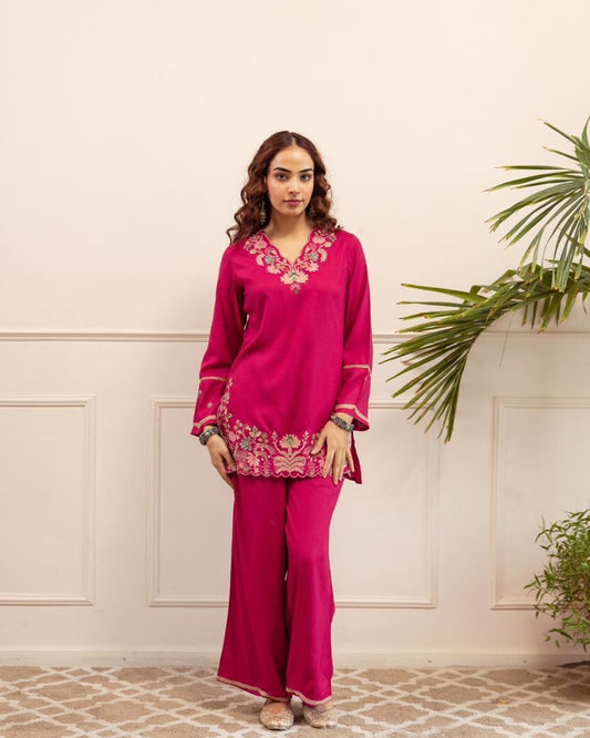 Woman wearing a bright pink traditional outfit with intricate designs against a light-colored wall.
