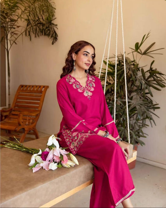 Woman in a pink traditional outfit sitting on a swing with plants and a wooden chair in the background.