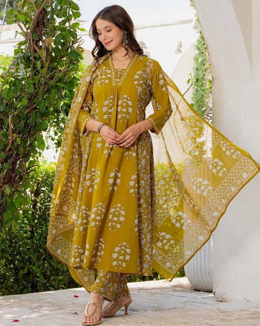 Woman in a yellow floral dress standing outdoors with greenery and a white architectural structure in the background.