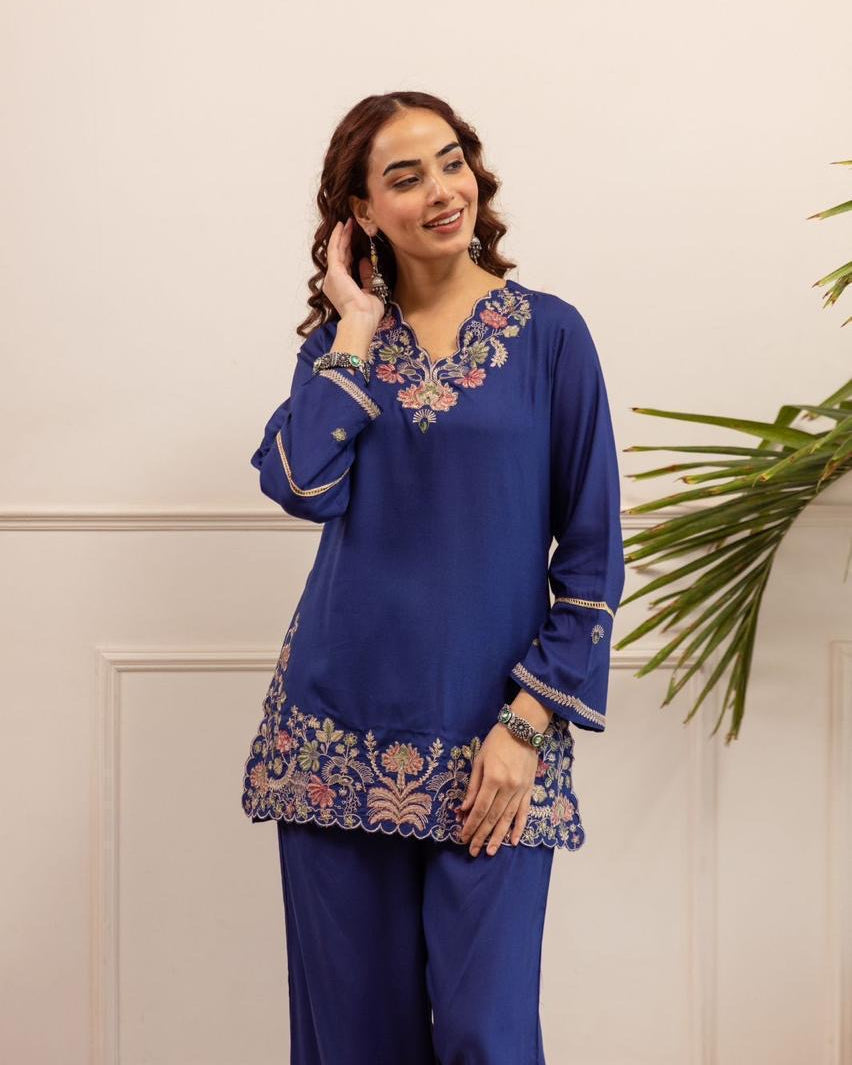 Woman wearing a blue embroidered outfit against a white wall with a plant.