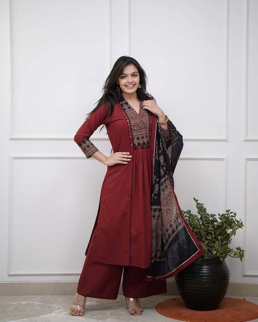 Woman in a red traditional outfit with a patterned scarf, standing against a white paneled wall.