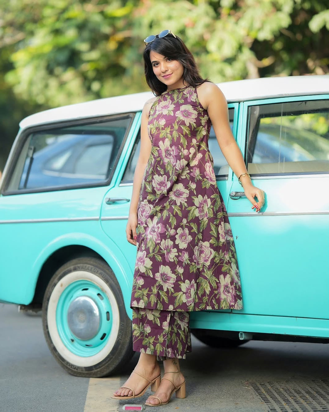 Woman in a floral dress standing next to a vintage turquoise car with greenery in the background
