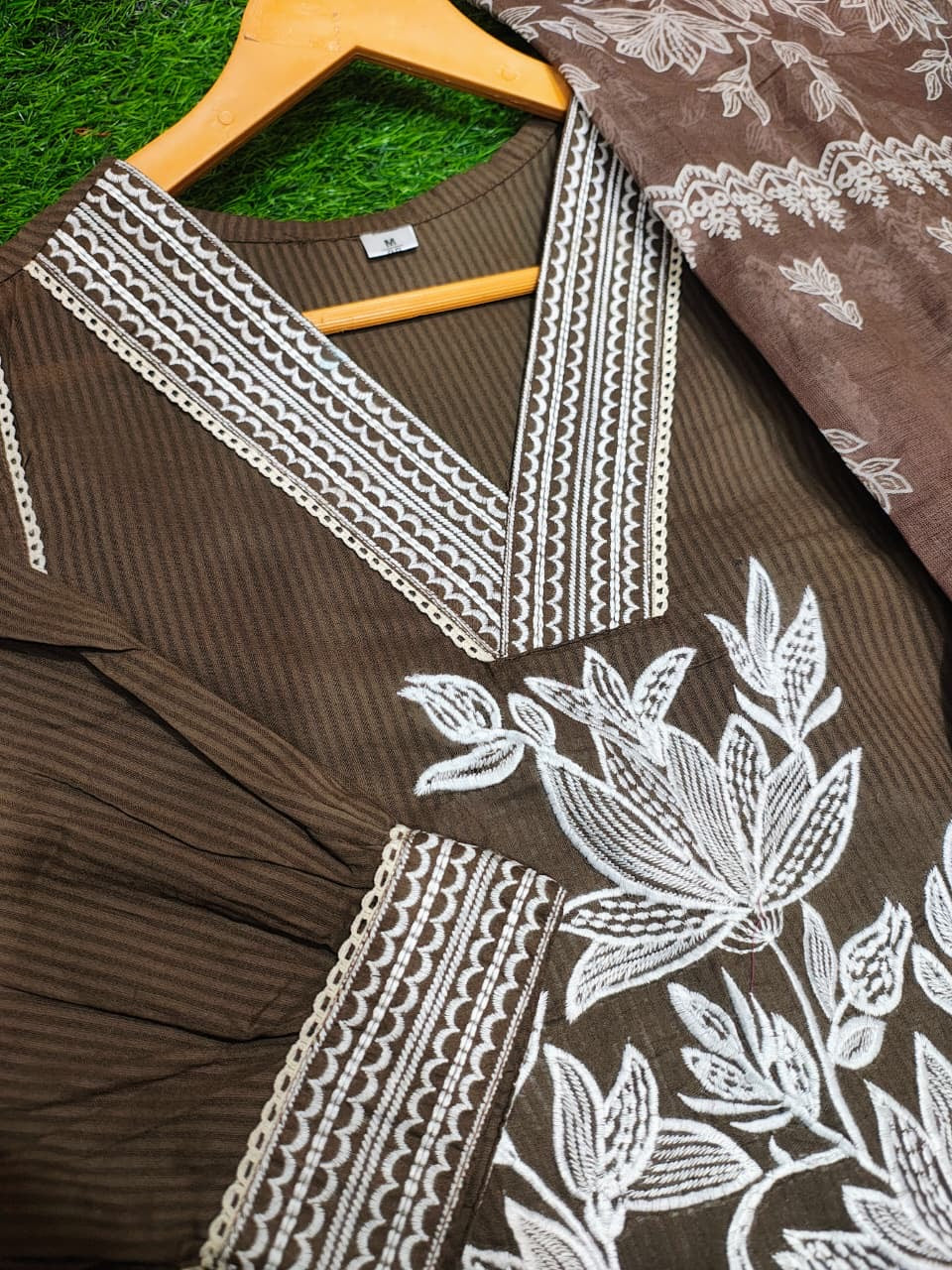 Brown garment with white floral embroidery on a hanger against a green grass background