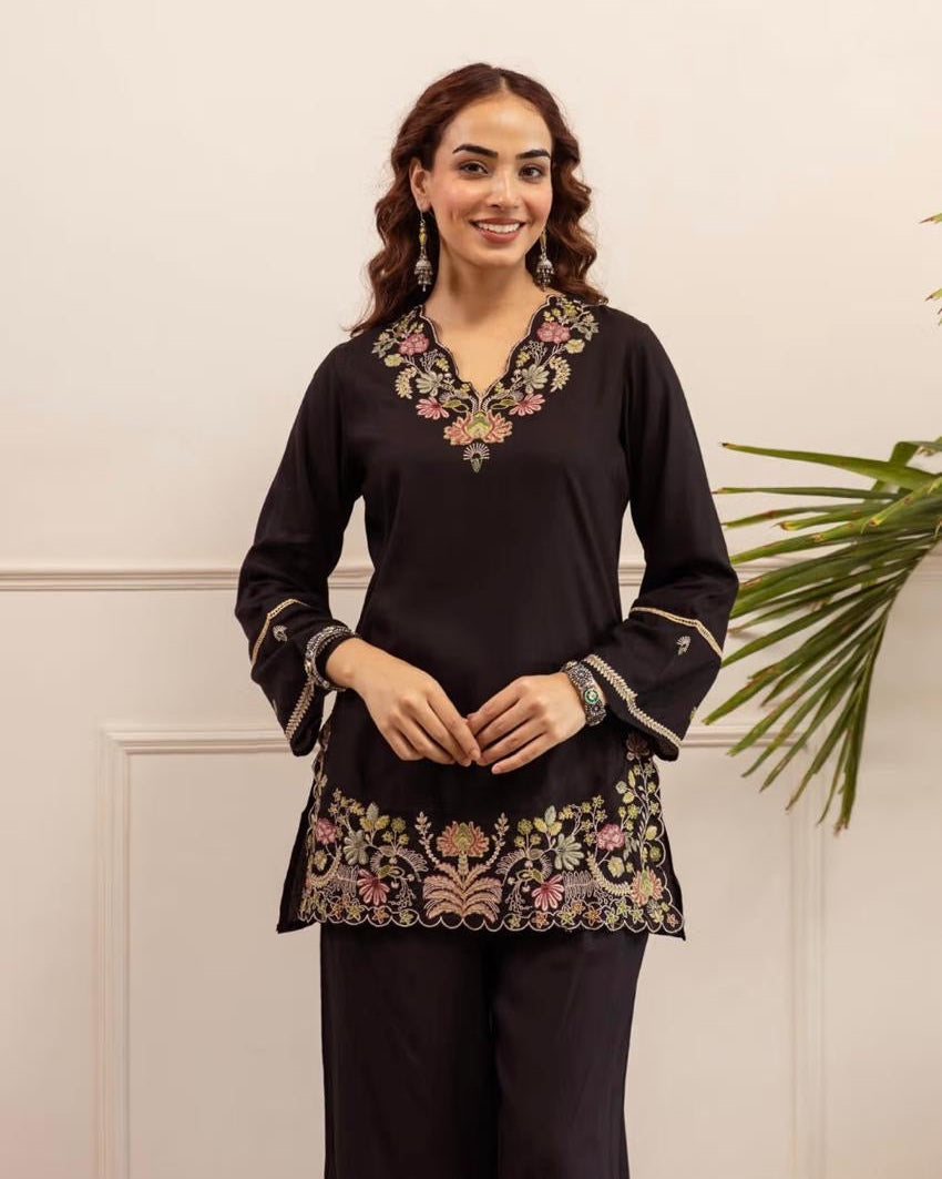 Woman wearing a black embroidered top with floral patterns against a white wall.