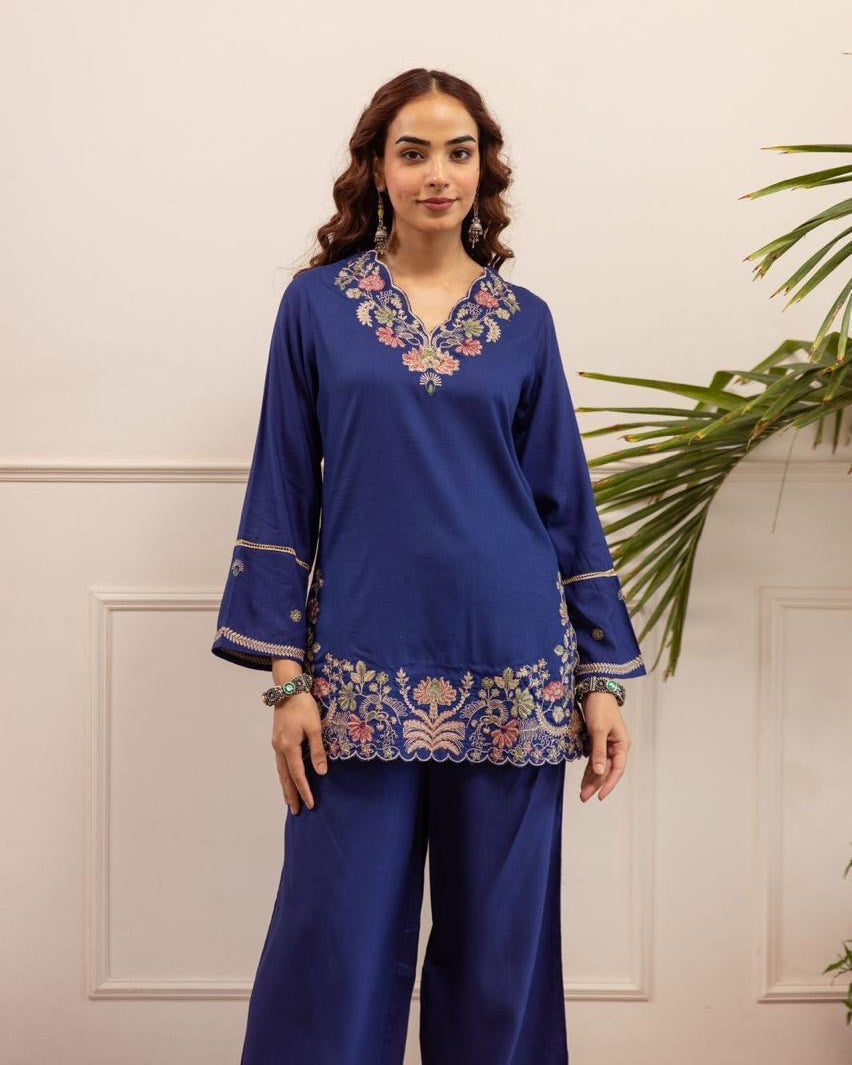 Woman wearing a blue embroidered outfit with a white wall and plant in the background