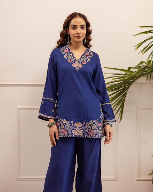 Woman wearing a blue embroidered outfit with a white wall and plant in the background