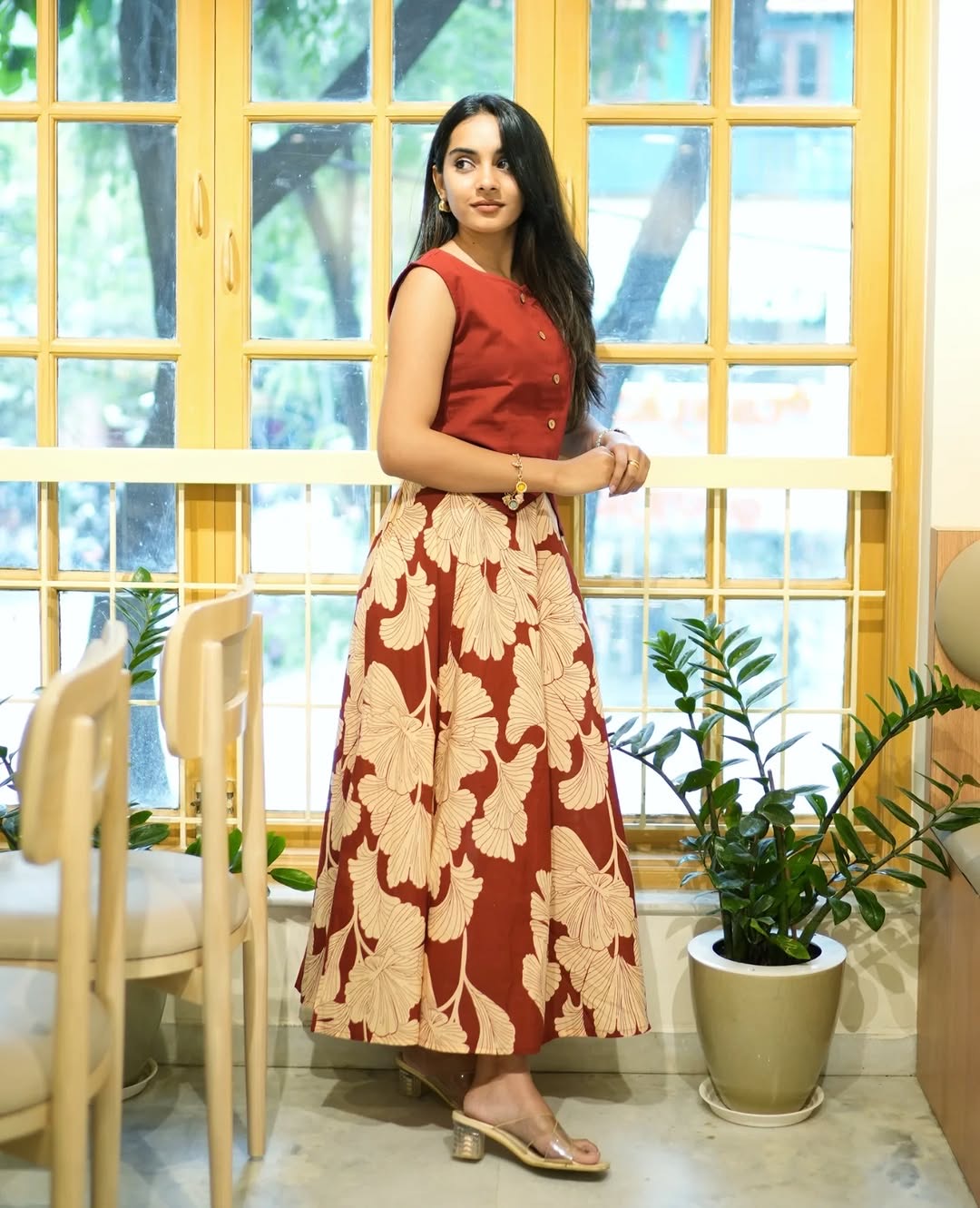 Woman in a red top and floral skirt standing in a room with large windows and plants.