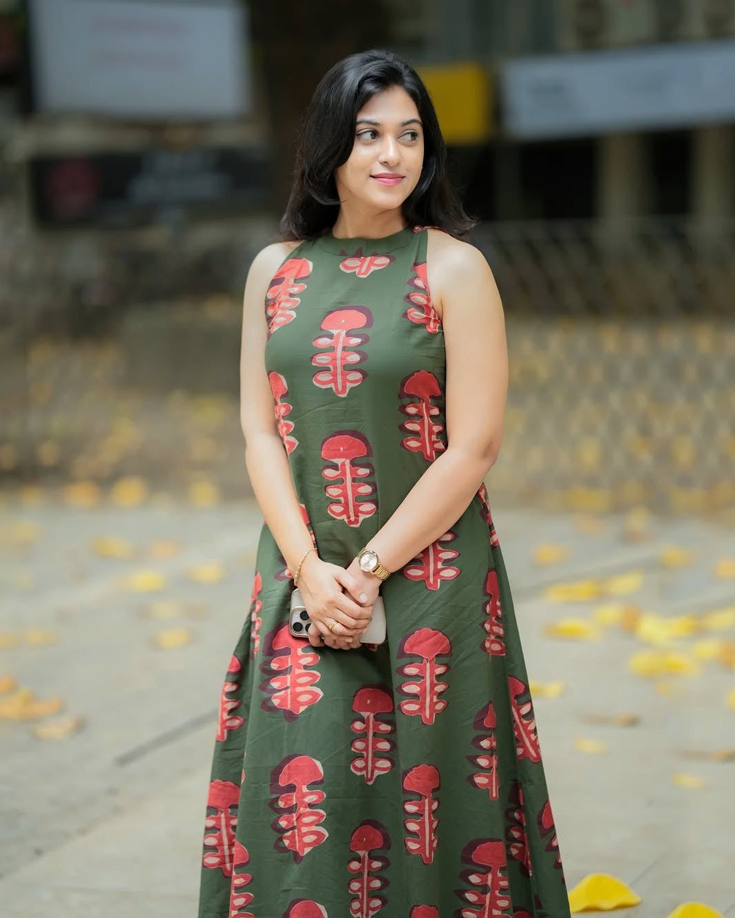 Woman wearing a green dress with red patterns standing outdoors on a leafy ground.