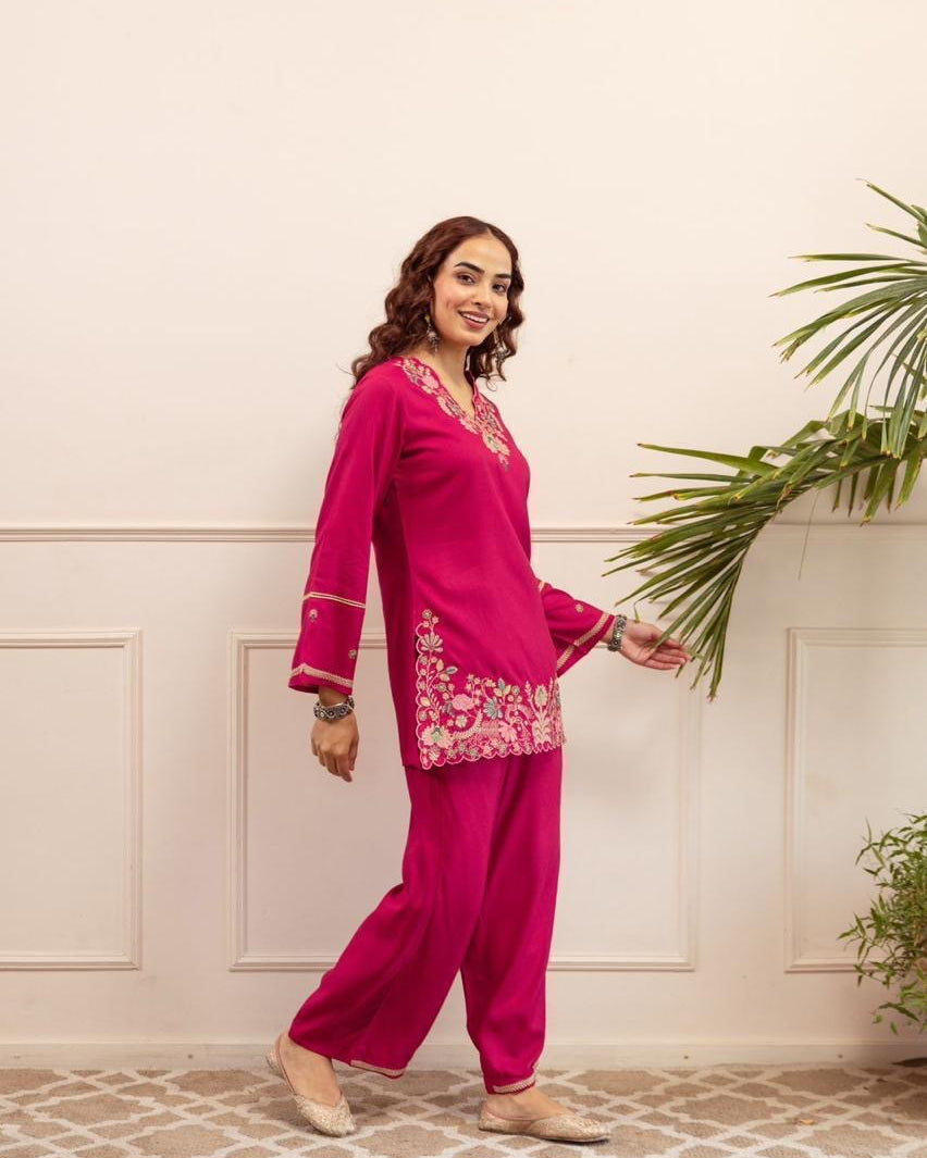 Woman in a pink traditional outfit standing against a light-colored wall with plants.