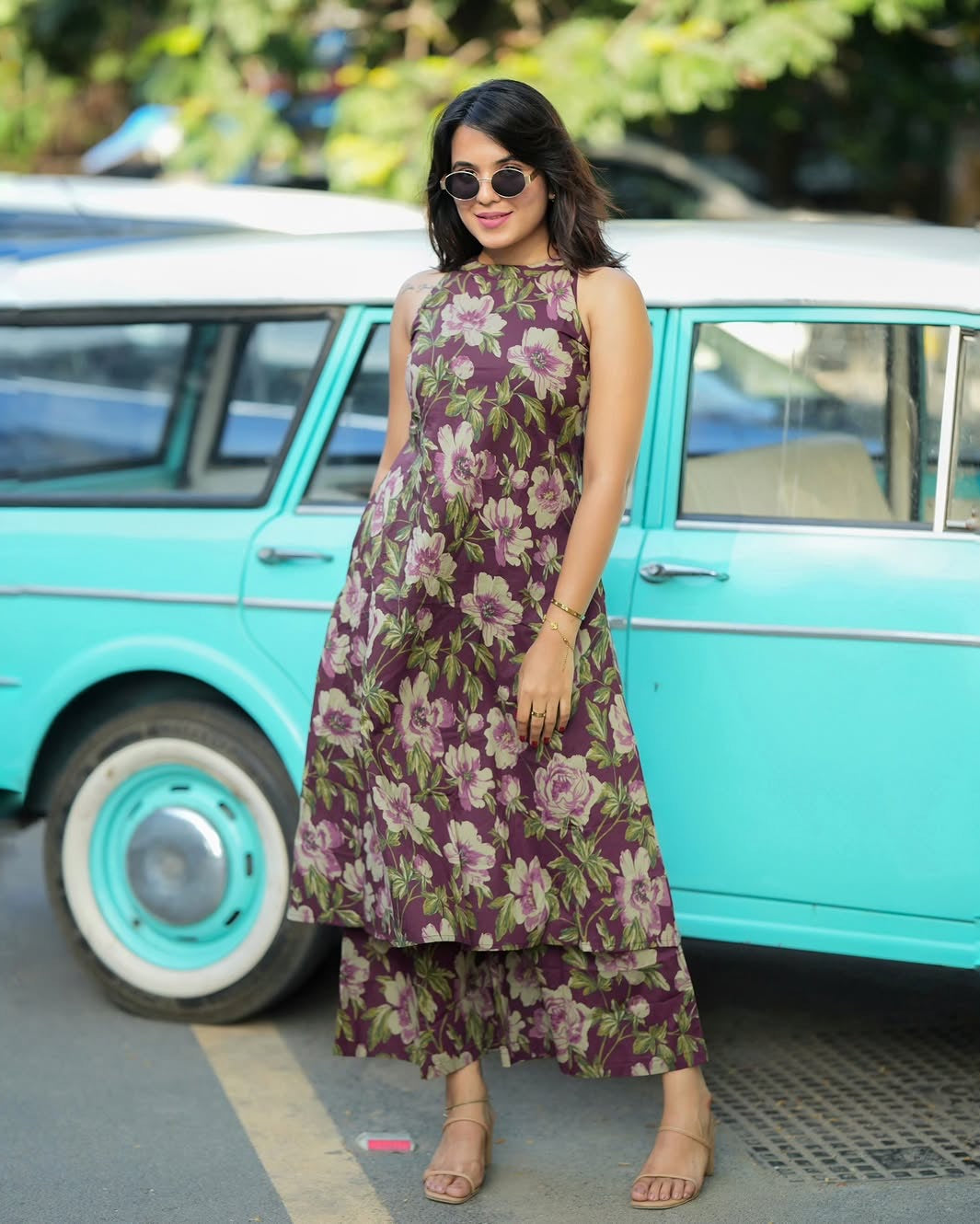 Woman in a floral dress standing in front of a turquoise vintage car.