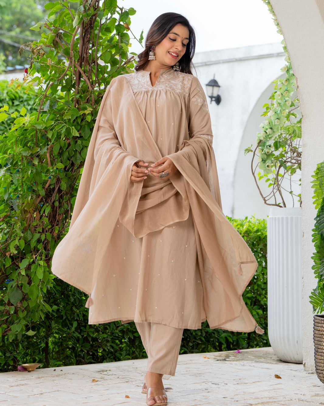 Woman in a beige traditional outfit standing outdoors with greenery and a white archway in the background.