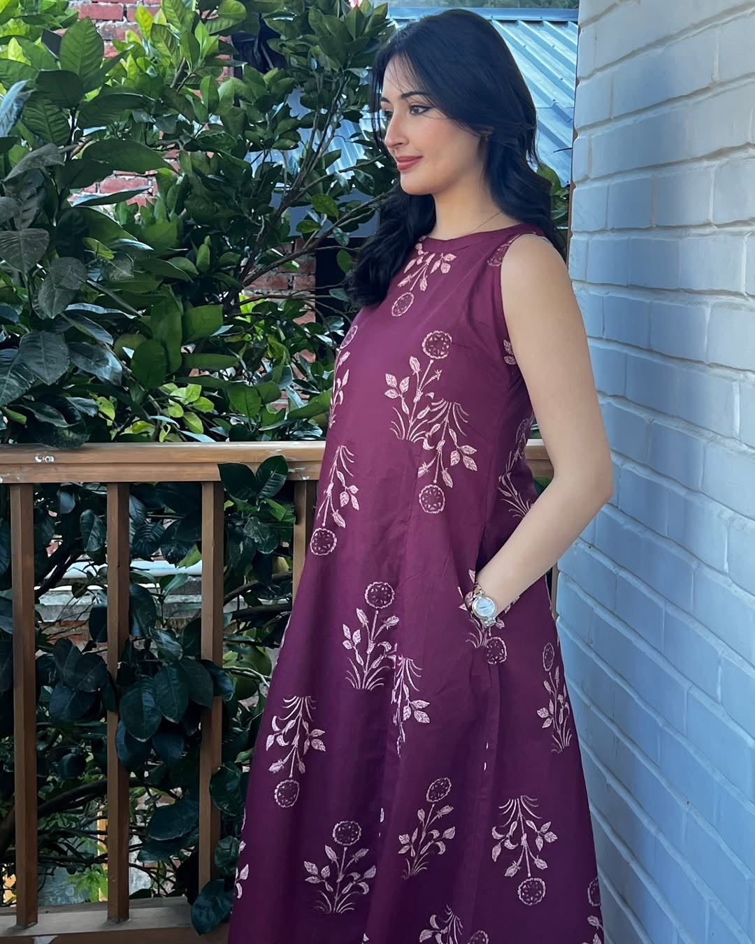 Woman in a purple dress with floral patterns standing against a white wall.