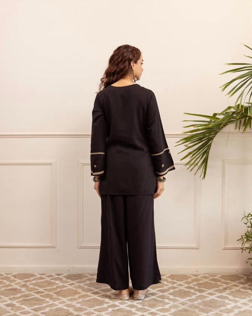 Woman wearing a black outfit with gold trim standing in a room with a decorative wall and plants.