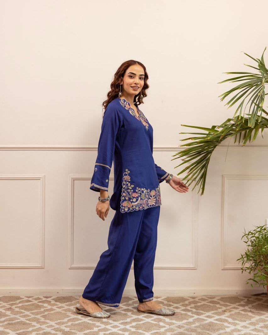 Woman in a blue traditional outfit standing in a room with a plant.