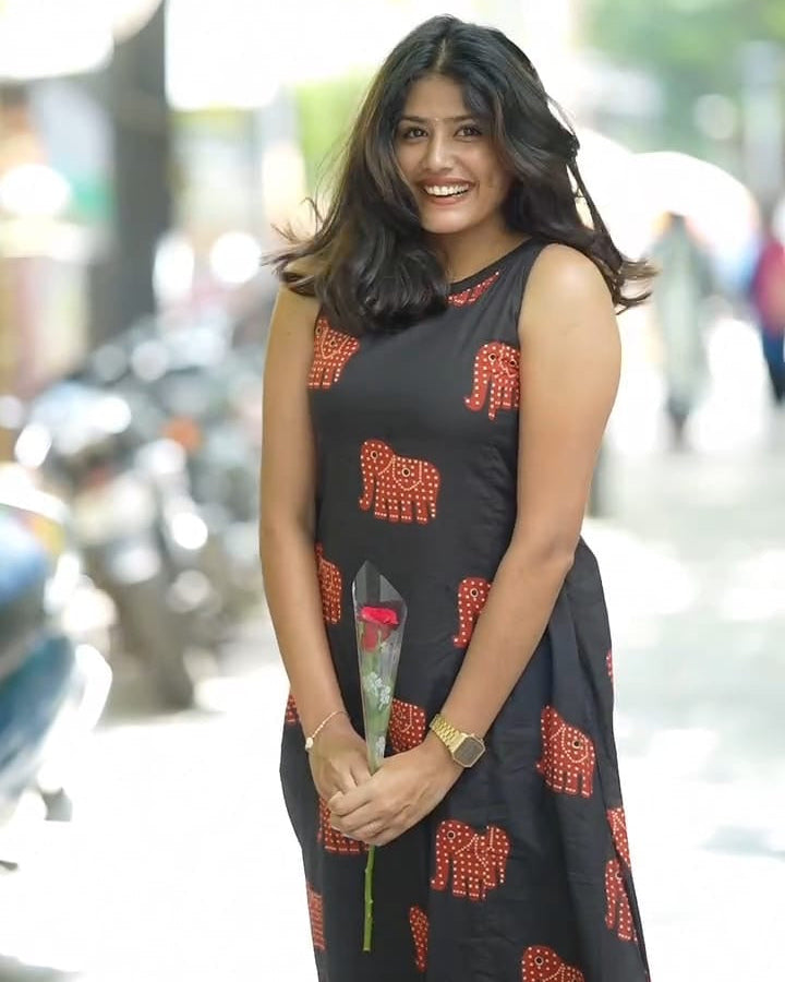 Woman in a black dress with red patterns holding a flower, standing outdoors.