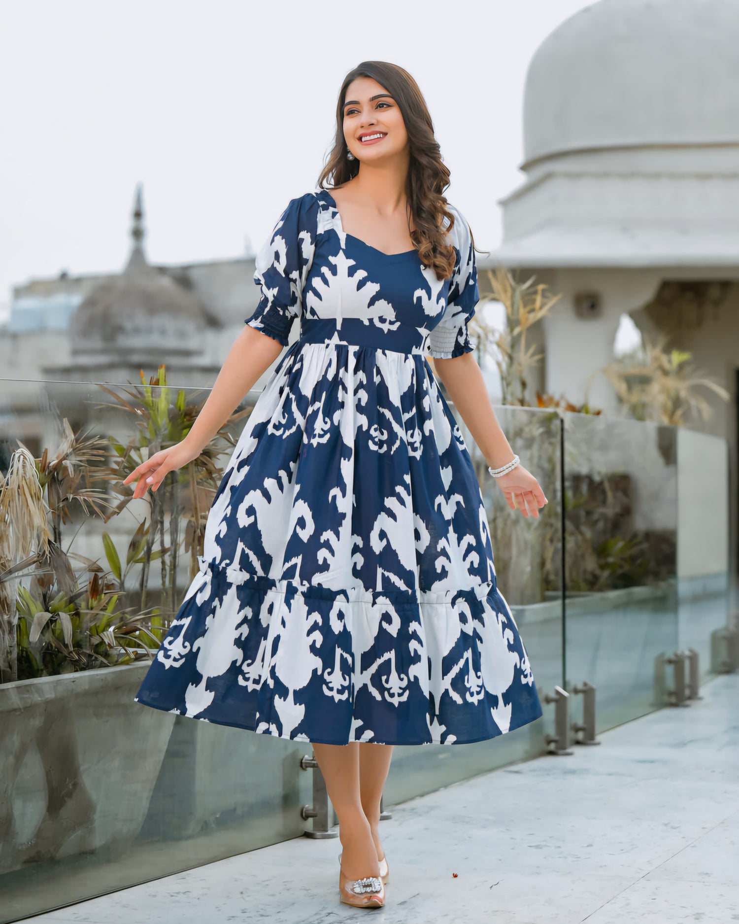 Woman in a blue and white patterned dress standing on a rooftop with plants and architectural elements in the background.