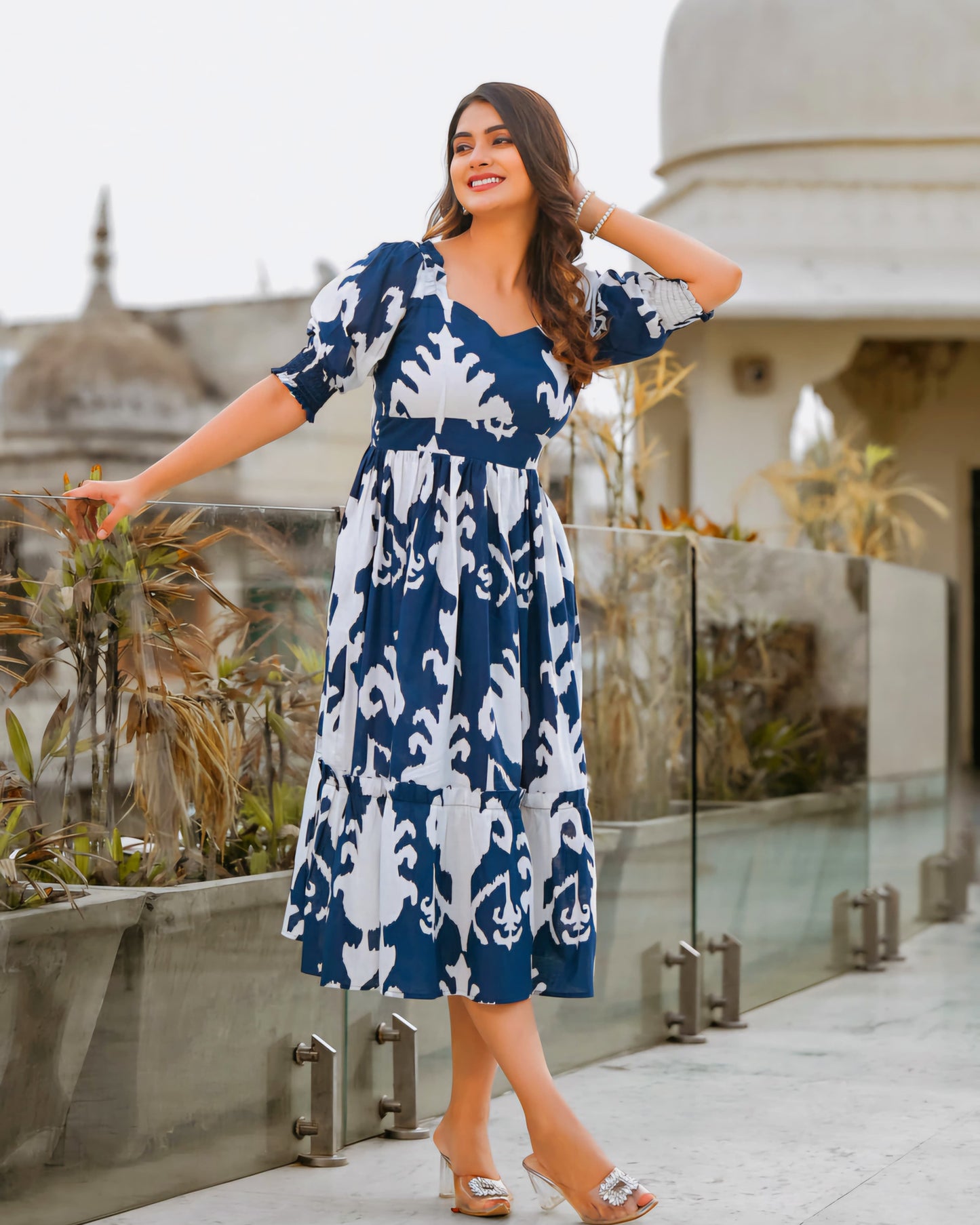 Woman in a blue and white patterned dress standing on a rooftop with architectural elements in the background.