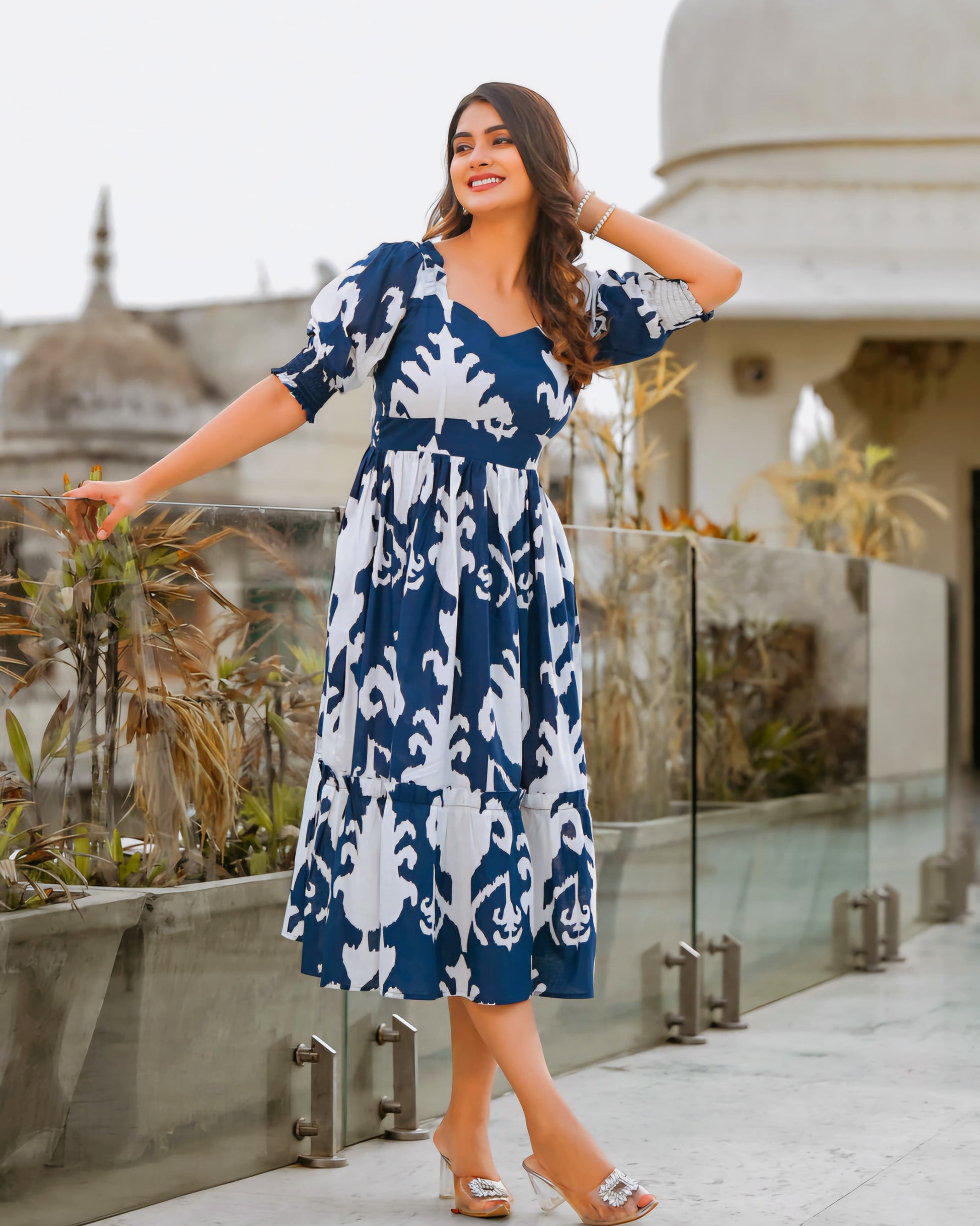 Woman in a blue and white patterned dress standing on a rooftop with architectural elements in the background.