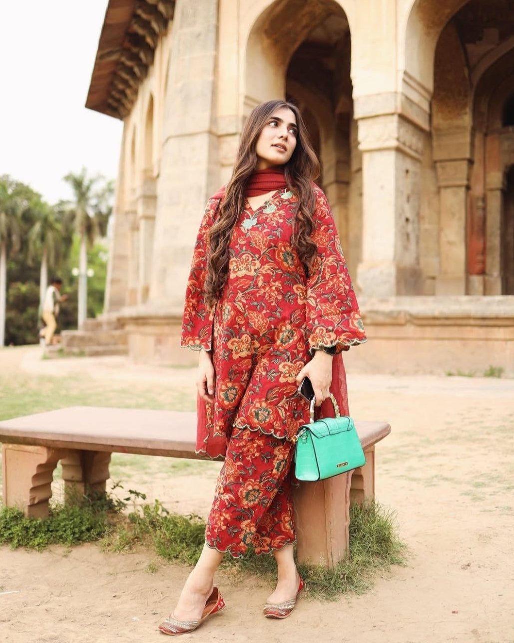 Woman in a red floral outfit holding a turquoise bag in front of historical architecture.