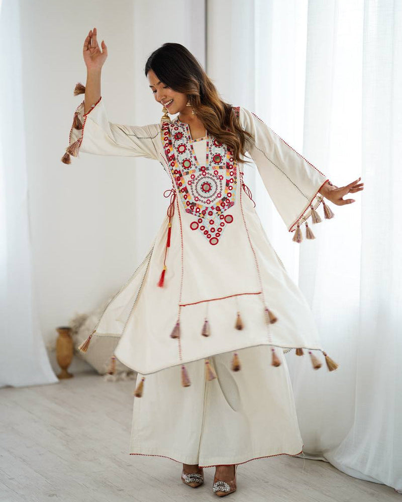 Woman in a white embroidered dress with red patterns and tassels, standing in a bright room.