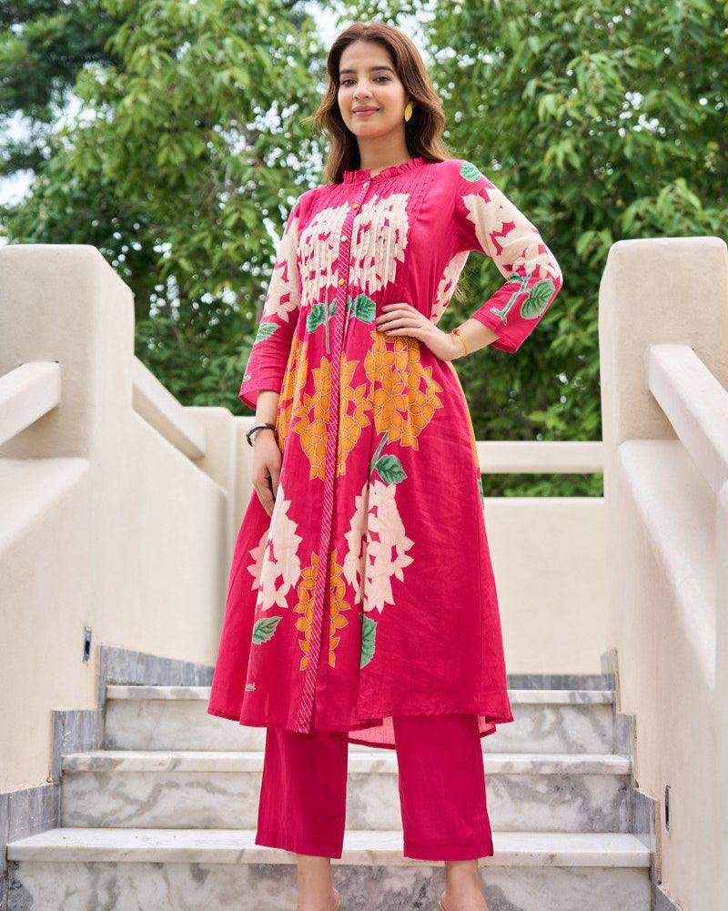 Woman in a pink embroidered outfit standing on steps with greenery in the background