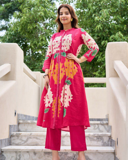 Woman in a pink embroidered outfit standing on steps with greenery in the background