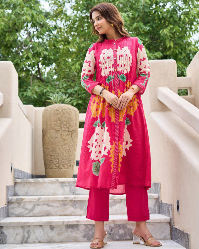 Woman in a bright pink traditional outfit with floral patterns standing on steps outdoors.