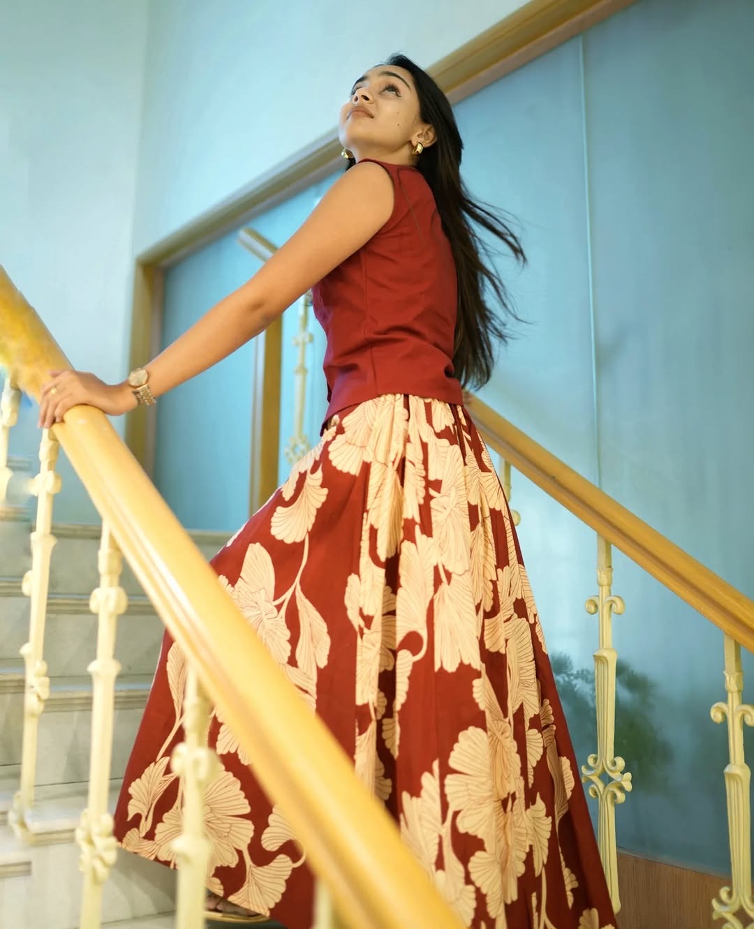 Woman in a red top and floral skirt standing on a staircase with a light blue wall in the background.