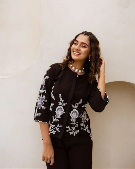 Woman wearing a black top with white floral embroidery against a beige wall.