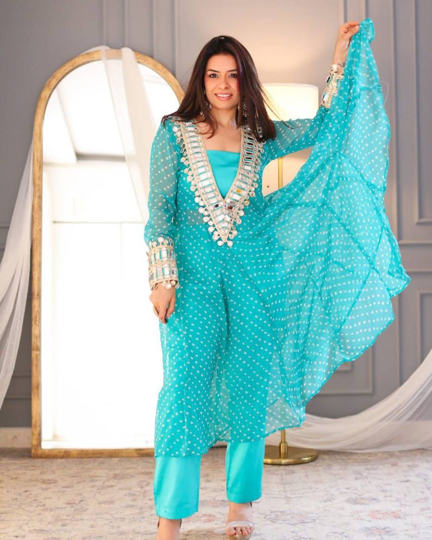 Woman in a turquoise traditional outfit with silver embellishments standing in a room with a large mirror.