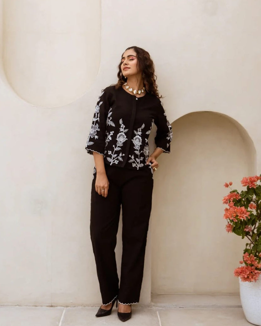 Woman in a black outfit with floral patterns standing against a white wall with a plant on the right.