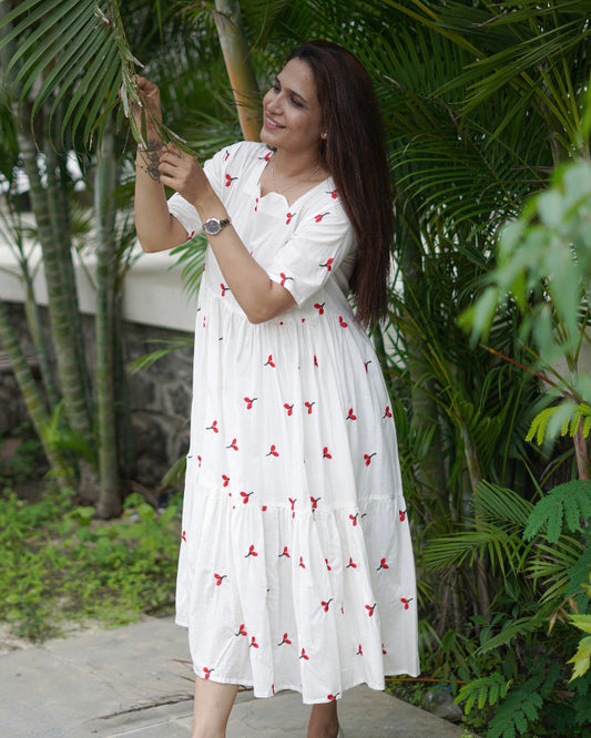 Woman in a white dress with red patterns taking a selfie outdoors among greenery.